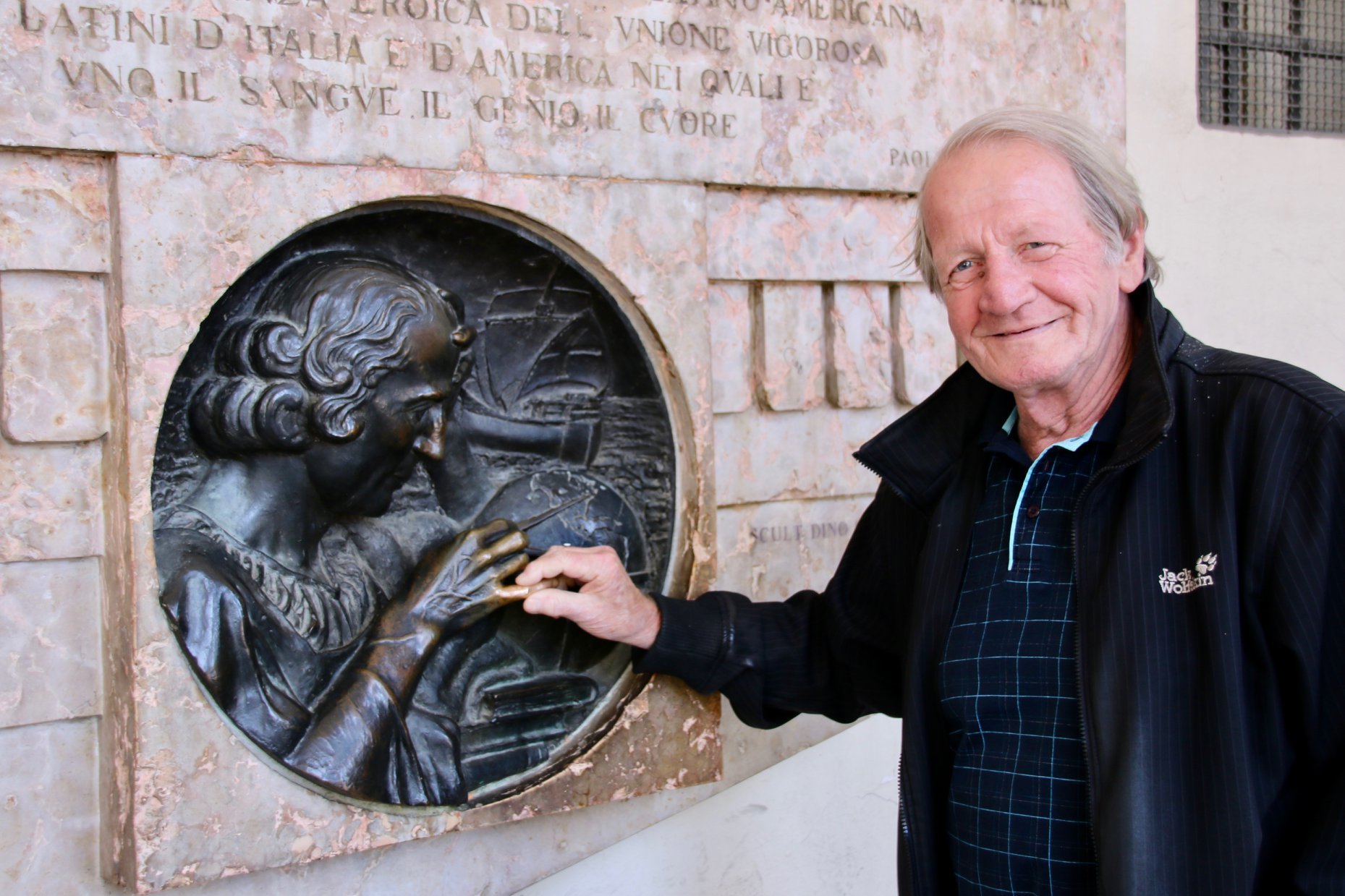 Herr Josef Wastler steht vor einer Wandskulptur und berührt den kleinen Fingerr 