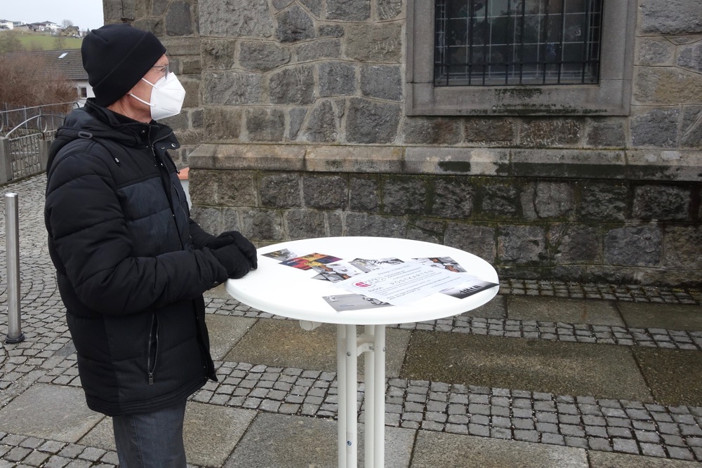 Ein Mann steht an einem Stehtisch am Kirchenvorplatz und bietet Postkarten an